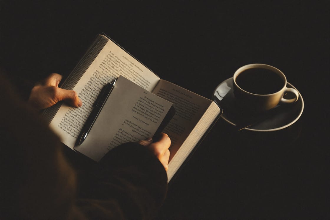 Hands holding an open book and a pen beside a cup of coffee in warm, low morning light.